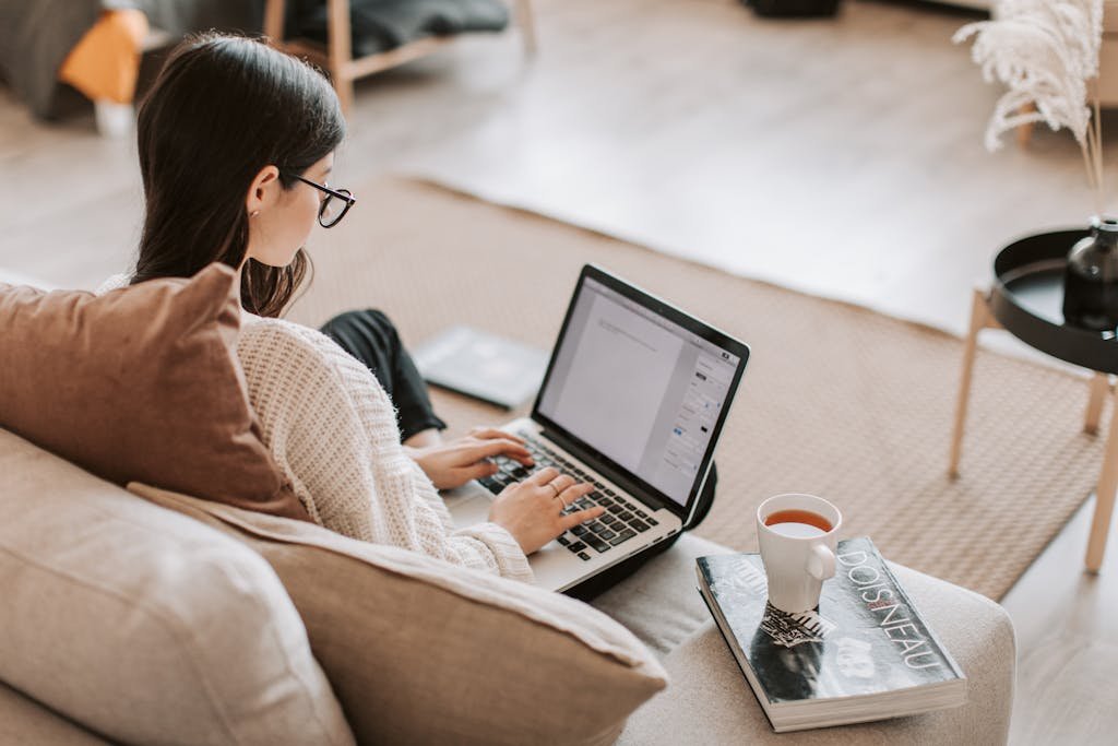 From above back view of full length female freelancer with crossed legs sitting on cozy sofa with cup of tea on book while typing text on laptop